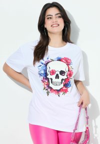 Camiseta blanca con un gráfico central de calavera adornado con patrones florales coloridos. El modelo la combina con leggings rosados brillantes y un bolso rosa.