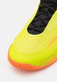 Neon gul atletik sko med tekstureret overflade, sort mesh tunge og orange sål. Har lagdelt design og fremtrædende syninger.