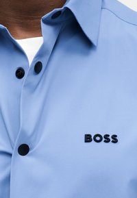 Gros plan d'une chemise bleu clair à col avec des boutons noirs et le logo "BOSS" sur la poitrine, portée sur un sous-vêtement blanc.