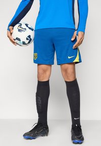 Μπλε ποδοσφαιρική φανέλα με ναυτικά μπλε λεπτομέρειες, μπλε σορτς με κίτρινο λογότυπο, μαύρες κάλτσες και μαύρα παπούτσια με μπλε λεπτομέρειες. Κρατάει μια λευκή ποδοσφαιρική μπάλα.