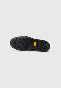 Suela de zapato de goma negra con un patrón de banda de rodadura robusto, detalle del logo amarillo, costura beige y múltiples puntos de tracción para una mejor adherencia.