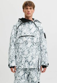 Jeune homme portant une veste technique à motifs marbrés blancs et gris et un pantalon assorti, se tenant contre un fond clair uniforme.