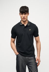 Άνδρας στέκεται με το πρόσωπο προς τα εμπρός φορώντας μαύρο πόλο με ριγέ γιακά και σκούρο γκρι παντελόνι, με το χέρι στην τσέπη, ουδέτερο φόντο.