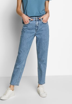 Osoba nosząca jasnoniebieskie jeansy z wysokim stanem, niebieski t-shirt włożony w spodnie, białe buty sportowe, stojąca z rękami w kieszeniach na jednolitym tle.