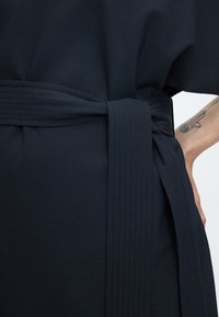 Κοντινό πλάνο μαύρης υφασμάτινης ζώνης δεμένης γύρω από τη μέση, με ορατή ραφή, και αντιβράχιο που δείχνει ένα χέρι και ένα τατουάζ.