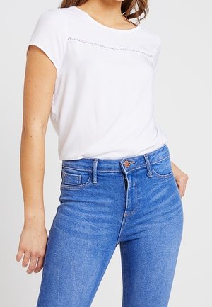 Bílý krátký rukáv top s texturovaným panelem přes hrudník, spárovaný s těsně přiléhajícími modrými džíny z denimu s klasickým zapínáním na knoflík a standardními kapsami.