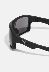 Schwarze Sportsonnenbrille mit dunklen Gläsern, gummiertem Rahmen und kantigem Design. Sie verfügt über ein metallisches Puma-Logo an der Seite und strukturierte Akzente.