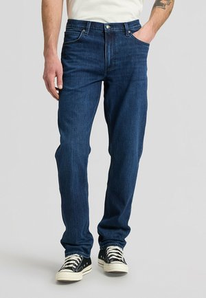Man wearing dark blue straight-leg jeans with black and white sneakers, standing with one hand in pocket against plain background.