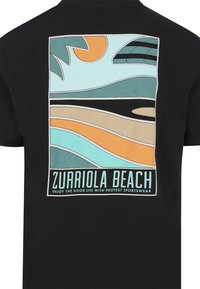Černé tričko s barevným grafickým potiskem vln, slunce a surfařské desky. Text zní "ZURRIOLA BEACH" a je doplněn geometrickými tvary.