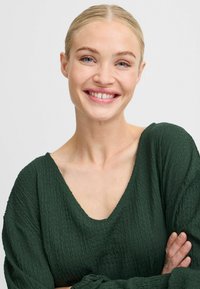 Tmavě zelená texturovaná blůza s hlubokým výstřihem do V a volnými rukávy, kterou nosí osoba s úsměvem a zkříženýma rukama. Jednoduché bílé pozadí.