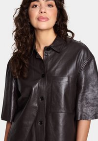 Brown leather shirt with a relaxed fit, short sleeves, a point collar, button closure, and a chest pocket. Smooth texture and subtle sheen.