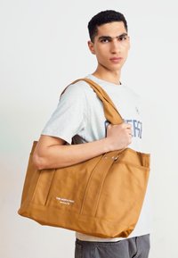 Tan tote bag with a smooth texture, featuring a spacious design, shoulder straps, and a logo on the front. Sturdy and functional.