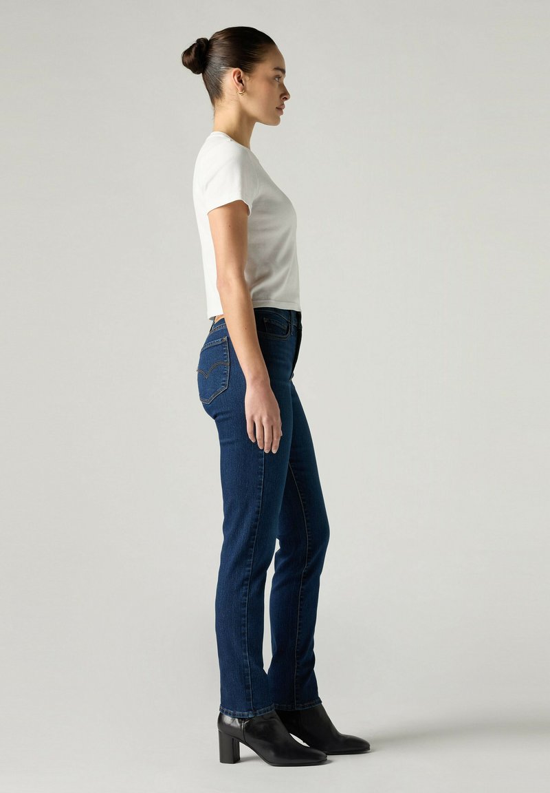 Femme avec une coiffure en chignon, portant un t-shirt blanc, un jean bleu foncé et des bottines noires à talons, debout de profil sur un fond uni.