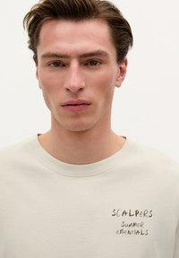 Helles beiges Sweatshirt mit Rundhalsausschnitt, das auf der linken Seite handgeschriebene Texte "SCALPERS SUMMER ESSENTIALS" zeigt. Sanfte Textur und lässige Passform.