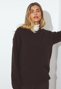 Pull en maille marron foncé à la coupe décontractée, avec un col rond, un ourlet et des poignets côtelés, porté par-dessus un haut de couleur claire.