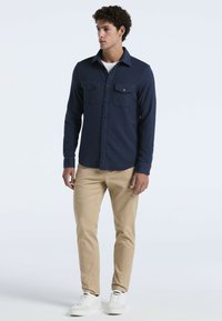 Jonge man staat gekleed in een marineblauw knoopshirt over een wit T-shirt, beige broek en witte sneakers, terwijl hij opzij kijkt tegen een effen achtergrond.