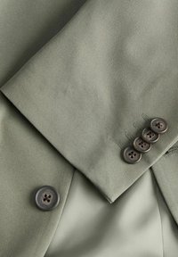 Μανίκι από σακάκι ανοιχτό πράσινο με τέσσερα σκούρα κουμπιά στη μανσέτα και ένα αντίστοιχο κουμπί μπροστά κοντά στην διπλωμένη πέτα.