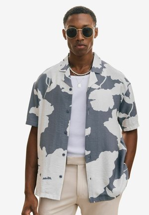 Mand iført runde solbriller, grå- og hvid blomsterkortærmet skjorte med knapper, hvid tanktop, beige bukser, med hænderne i lommerne.