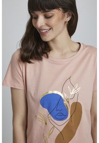 T-shirt en coton rose avec un imprimé graphique abstrait comprenant des formes bleues, dorées et brunes, y compris des éléments de visage et de feuille. Manches courtes.