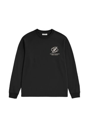 Mand iført en sort sweatshirt med hvid tekst på brystet og en brun kasket, der kigger til siden, hånd i lommen på mørkeblå jeans.