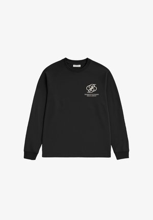 Mand iført en sort sweatshirt med hvid tekst på brystet og en brun kasket, der kigger til siden, hånd i lommen på mørkeblå jeans.