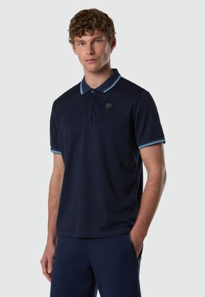 Junger Mann mit lockigem Haar, der ein marineblaues Poloshirt mit hellblauer Einfassung am Kragen und an den Ärmeln trägt und mit einer Hand in der Tasche steht.