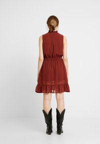 Vestido sin mangas color marrón con dobladillo con volantes, cinturilla elástica y cierre en la espalda con abertura, combinado con botas de cowboy negras.