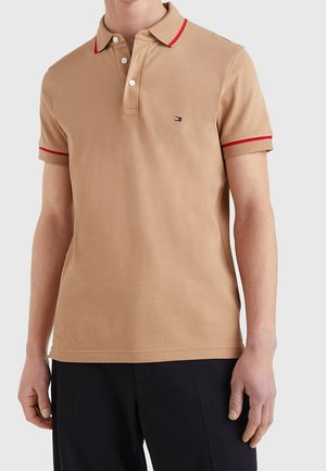 Beige kurzärmeliges Poloshirt mit rotem Besatz am Kragen und an den Manschetten, weißen Knöpfen und kleinem Logo auf der linken Brust, getragen mit schwarzer Hose.