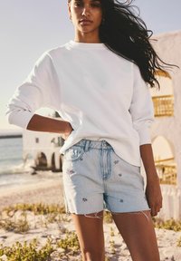 Sweat-shirt blanc à manches longues associé à un short en denim bleu clair. Le short présente des broderies florales et des bords effilochés. Fond de plage.