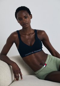 Mornarska sportska grudnjak s bijelim logotipom "TOMMY HILFIGER", s V izrezom. Zelene kratke hlače s logotipom na pojasu koji se podudara. Glatka tkanina, minimalistički dizajn.