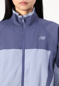 Žena v dvoubarevném fialovém zipovém saka New Balance s vysokým límcem, zachycena od ramen k rtům na bílém pozadí.