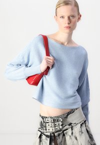 Lys blå ribbestrikket genser med brede ermer, kombinert med et grått skjørt med tie-dye mønster og knappdetaljer. Modellen holder en rød håndveske.