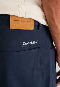 Navy blue pants with embroidered "Prohibited" text above the back pocket and a tan leather patch also labeled "PROHIBITED."