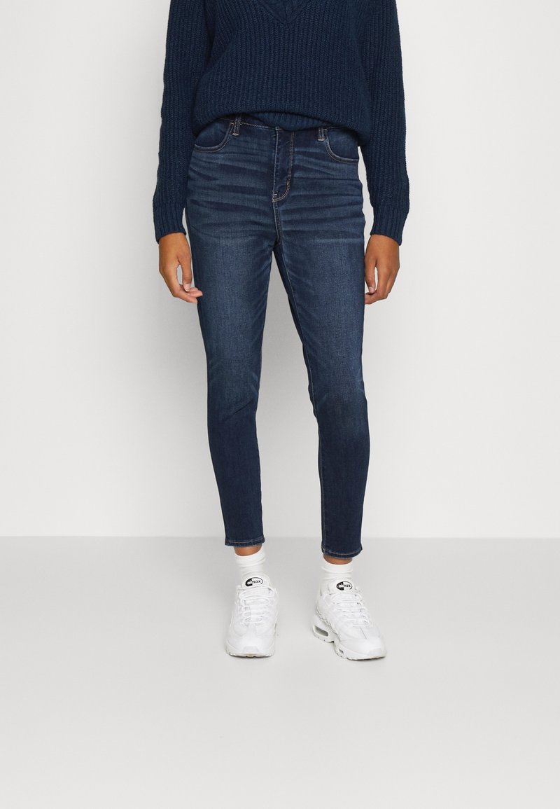 Jeans slim en denim bleu foncé, à taille mi-haute, avec un design classique à cinq poches et un léger délavage. Associés à des baskets blanches.