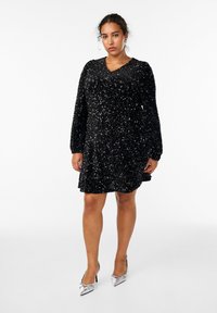 Abito nero di paillettes con scollatura a V, maniche lunghe e gonna svasata. Scarpe con tacco alto argentate completano il look.