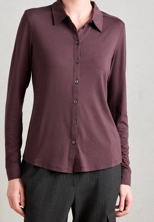 Kobieta ubrana w bordową koszulę na guziki z długim rękawem oraz ciemne spodnie w cienkie prążki, stojąca na jednolitym jasnym tle.