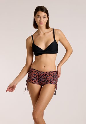 Naine, kellel on musta värvi bikiinid ja pruunide leopardimustriliste küljepaeltega ujumispüksid, seisab neutraalse tausta ees.