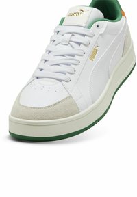 Zapatilla blanca con acentos verdes y punta de gamuza. Presenta cordones blancos, un cuello acolchado y un logo dorado en la lengüeta. Suela gruesa con fondo verde.