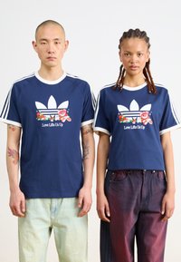 Marineblå t-skjorter med hvit kant og Adidas-logo; grafiske blomster og teksten "Kjærlighet løfter oss opp" foran. Modellen har på seg grønne og burgunder bukser.
