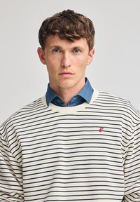 Kremowy sweter w paski z granatowymi liniami, o oversize'owym kroju, z prążkowanym kołnierzem, noszony z dżinsową koszulą. Posiada małe czerwone logo na piersi.