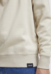 Sudadera color crema hecha de tela suave, con puños y dobladillo acanalados, y una etiqueta negra que muestra "!SOLID" en la parte inferior izquierda.