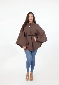 Capa grande marrón con un cuello alto, que cuenta con un solo botón y un cinturón negro. Se lleva con jeans skinny azules y tacones transparentes.