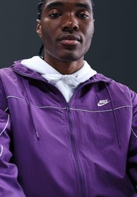 Giacca Nike viola con accenti bianchi, chiusura zip davanti e cappuccio integrato, indossata sopra un felpa bianca. Texture liscia, design sportivo.