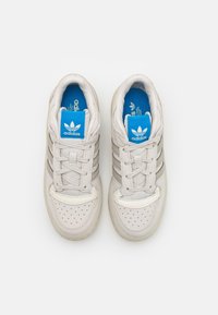 Bílé kožené tenisky se třemi stříbrnými pruhy, perforovanou špičkou a modrým logem Adidas na jazyku. Texturovaná gumová podrážka.