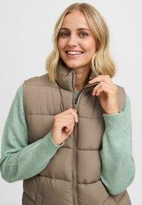 Gilet rembourré beige avec col montant, fermeture éclair et poches latérales, superposé à un pull léger vert clair avec des poignets côtelés.