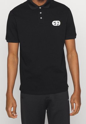 Man draagt een zwart polohemd met korte mouwen, drie knopen en een wit geborduurd logo op de linkerborst.