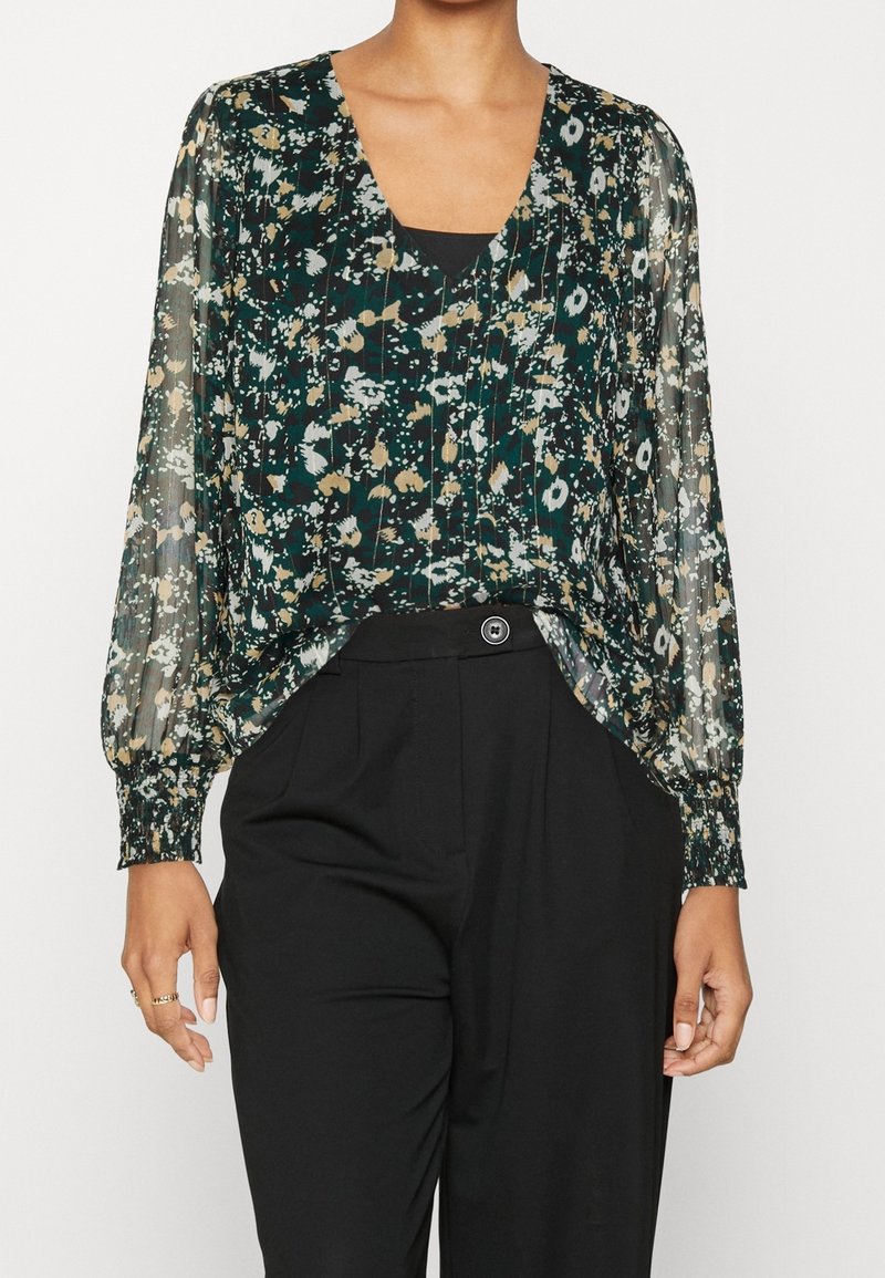 Femme portant un chemisier transparent à motif floral avec des manches longues bouffantes, rentré dans un pantalon noir taille haute.