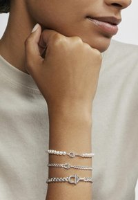 Tres pulseras: una con perlas, dos en cadenas de plata, con acentos de hardware distintos. Llevadas en una muñeca contra un fondo de tela suave.