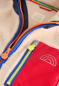 Kurtka polarowa z kremowym wnętrzem, niebieskim zewnętrznym materiałem z pomarańczowym zamkiem błyskawicznym, czerwonym kieszonką z żółtym zamkiem błyskawicznym oraz detalem z logo w kształcie tęczy.