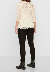 Lace top in cream with three-quarter sleeves, scalloped hem, worn with black slim-fit pants and ankle boots. Back view, plain background.